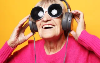 Cheerful old woman wearing pink sweater and big sunglasses listening to music with headphones over yellow background