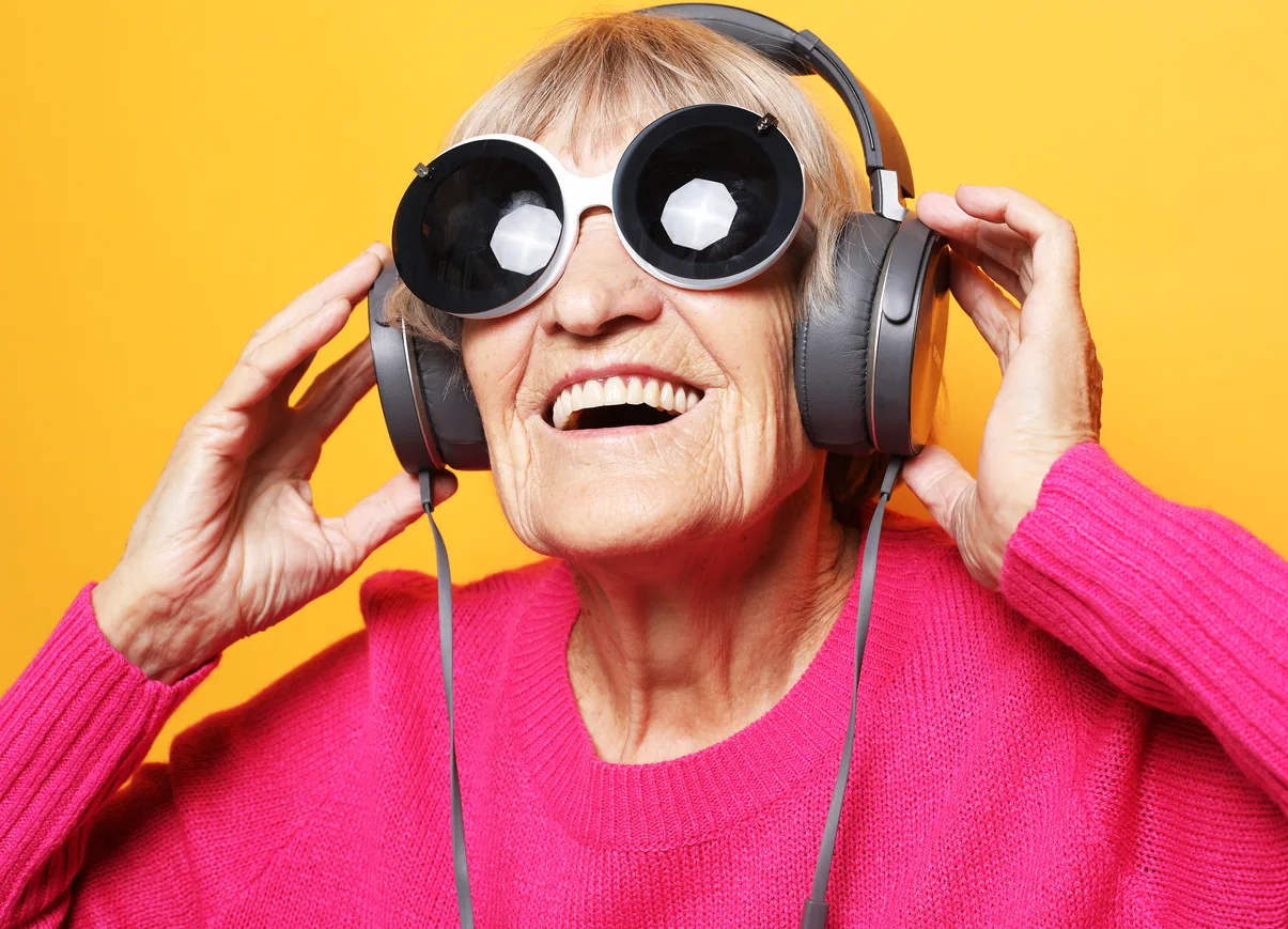 Cheerful old woman wearing pink sweater and big sunglasses listening to music with headphones over yellow background